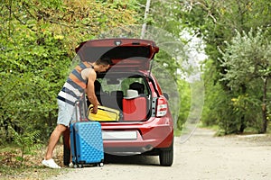 Happy man packing suitcases into car trunk near forest