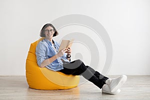 Happy man reading book and smiling at camera while sitting in comfortable beanbag chair over light wall, copy space