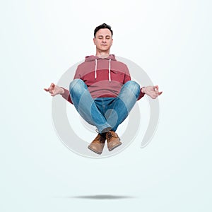 Happy man in red sweatshirt and jeans closing eyes relaxed levitates in air in lotus position, on light blue background