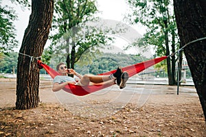 Happy man resting on a lounger on a hammock in the park and calling on the phone against the backdrop of the cityscape with the