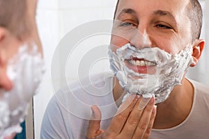 Happy man shaving in front of a mirror in the bathroom. Hygiene, skin care.