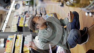 Happy man in shirt sitting near a cart in supermarket choosing groceries. Vertical footage. Making a squat to take from