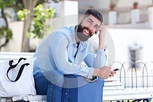 Happy man sitting outside with suitcase and mobile phone