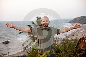 Happy man standing with arms wide open on green hill in front of seashore against seascape and enjoying vacation.