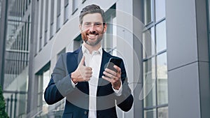 Happy man standing outdoors holding phone smiling businessman using new mobile app for business tasks showing thumb up