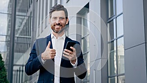 Happy man standing outdoors holding phone smiling businessman using new mobile app for business tasks showing thumb up