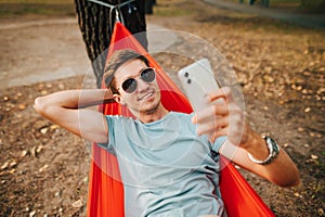 Happy man in sunglasses lies in a hammock on a tree in the park and takes a selfie on a smartphone with a smile on his face.