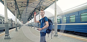 Happy man tourist with backpack stand on railway station platform, greeting friends or saying goodbye, waving his hand. Travel by