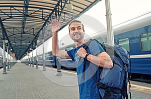 Happy man tourist with backpack stand on railway station platform, greeting friends or saying goodbye, waving his hand. Travel by photo