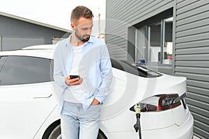 Happy man using smart phone and charging car at vehicle charging station