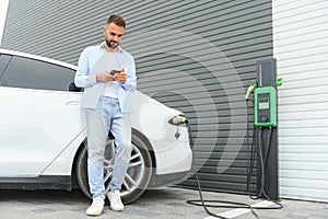 Happy man using smart phone and charging car at vehicle charging station