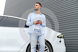 Happy man using smart phone and charging car at vehicle charging station