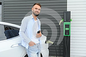 Happy man using smart phone and charging car at vehicle charging station