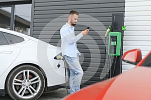 Happy man using smart phone and charging car at vehicle charging station