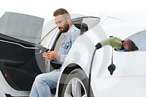 Happy man using smart phone and charging car at vehicle charging station