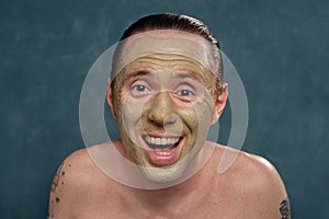 Happy man with wet long hair in green clay mask on his face waiting of spa procedures