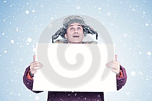 A happy man in winter clothes holds a white empty banner in his hands and looks at the sky with his mouth open.