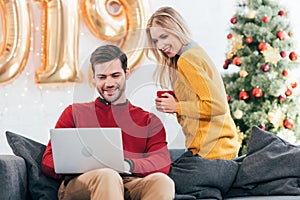 happy man and woman with cup of coffee using laptop together at home with 2019 golden balloons