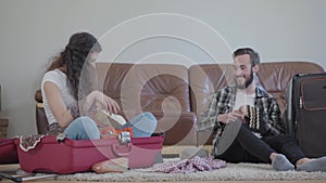 Happy man and woman on the floor at home in front of leather sofa, packing a suitcase before travel. The wife throwing