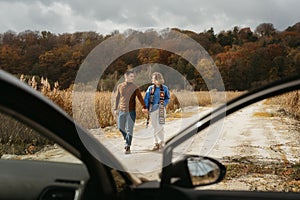 Happy man and woman are walking down a country road next to their car holding hands, adult couple on road trip in autumn