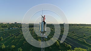 Happy man at work on top of cellular antenna, technician on radio telecommunication tower raises hand with thumb-up on