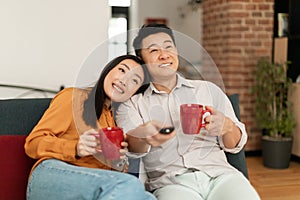 Happy married asian couple spending time at home together, drinking coffee and watching tv, relaxing on couch