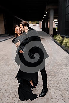 Happy married couple dancing at the streets, hugging, wearing stylish black looks. Handsome joyful beard man is holding