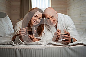 Happy married couple enjoying caffeinated drink in bedroom after awakening