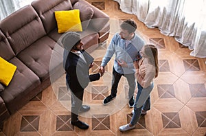 Cheerful married couple and friendly realtor shaking hands after making real estate deal indoors, above view