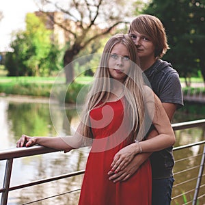 A happy married couple Husband hugs a beautiful adult pregnant wife. walks in a park by the lake. Second trimester.