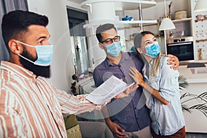 Happy married couple in medical masks standing in the new house with male real-estate agent. Pandemic concept