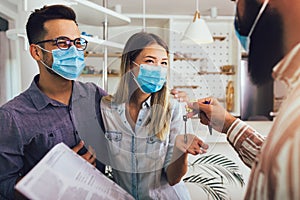 Happy married couple in medical masks standing in the new house with male real-estate agent. Pandemic concept