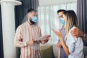 Happy married couple in medical masks standing in the new house with male real-estate agent. Pandemic concept