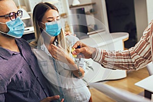 Happy married couple in medical masks standing in the new house with male real-estate agent. Pandemic concept