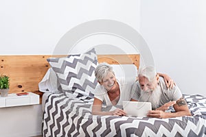 Happy married senior couple laughing together sitting in bed watching a fun video on tablet - Mature people having fun -