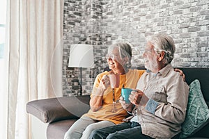 Happy mature 50s husband and wife sit rest on comfortable sofa in living room enjoy tea talking, smiling elderly 60s couple relax