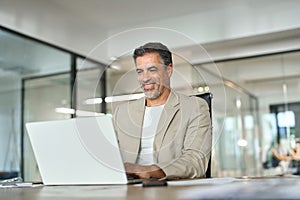 Happy mature bank manager looking at laptop in office sitting at desk.