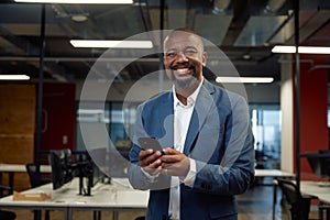 Happy mature black man in businesswear looking at camera while using mobile phone in office