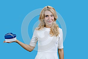 Happy mature blonde woman holding blue heart shape gift box on her right palm.