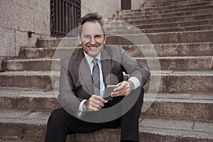 Happy mature business entrepreneur man sitting outdoors urban stairs working and using smart phone