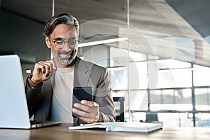 Happy mature business man sitting at office desk using mobile phone. Copy space