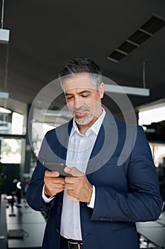 Happy mature business man wearing suit standing in office using mobile phone.