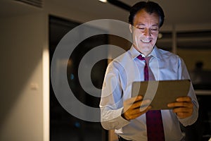 Happy Mature Businessman In Office At Night Using Digital Tablet Computer