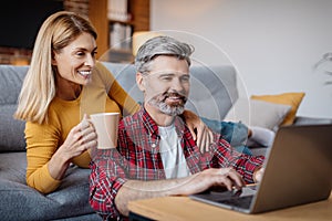 Happy mature caucasian wife and husband looking at laptop, enjoy coffee break in free time in living room