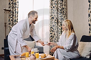 happy mature couple in bathrobes smiling each other while having breakfast together