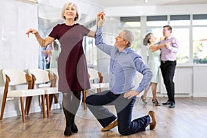 Happy mature couple enjoying retro ballroom dancing in dance salon, man standing on knee and holding hand of woman