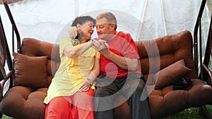 Happy mature couple man and woman swinging on swing in yard in rain, talking, kissing, having fun and fooling around