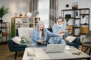 Happy mature family playing video games at home, sitting on sofa with laptop and using joysticks.