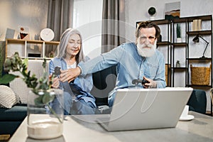 Happy mature family playing video games at home, sitting on sofa with laptop and using joysticks.