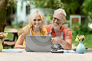 Happy Mature Farmers Couple Using Laptop Together Outdoors In Their Garden